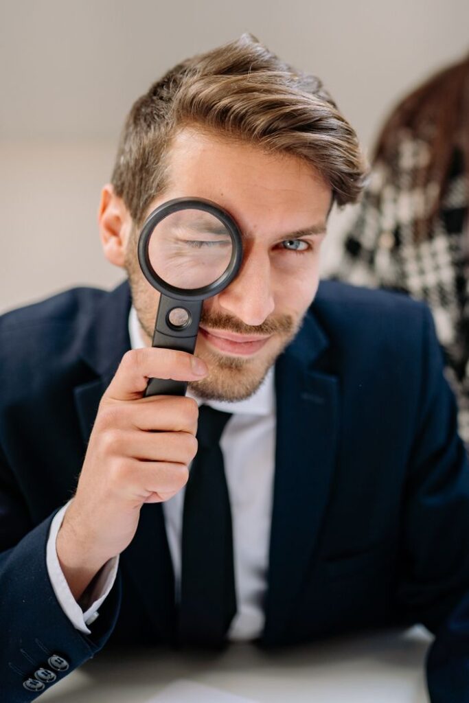 Young man in a suit smiling while holding a magnifying glass indoors.